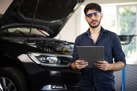 Portrait of caucasian bearded handsome man auto mechanic or manager in goggles smiling to camera in garage. Cheerful guy standing at cars service.