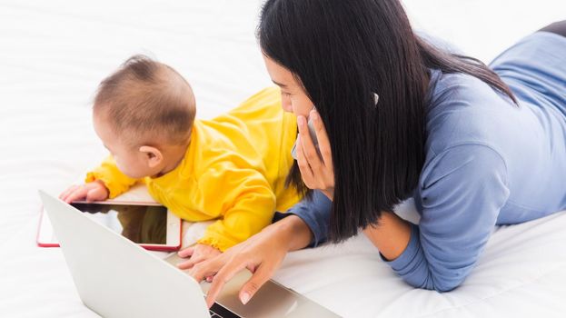 Asian young mother work from home busy with her baby daughter on laptop computer and she calling smartphone for communication with the customer, Freelance career for a young mom