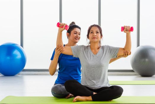 Asian young woman teacher teach adult student doing yoga hands holding dumbbells training body in studio. Two woman exercise workout with dumbbells, female sport fit workout concept