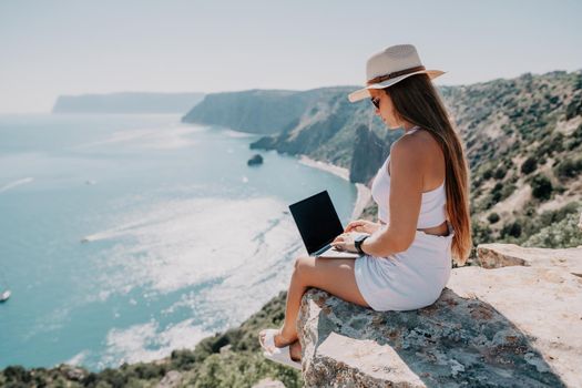 Successful business woman in yellow hat working on laptop by the sea. Pretty lady typing on computer at summer day outdoors. Freelance, travel and holidays concept.