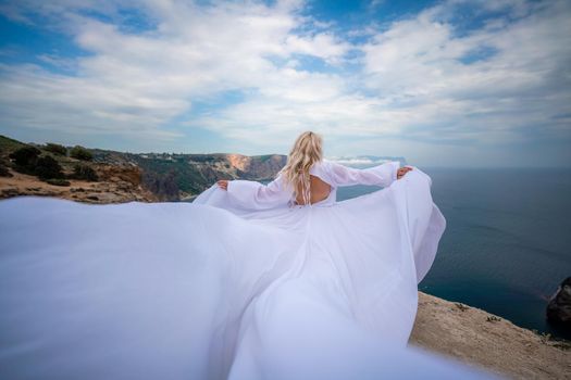 Blonde with long hair on a sunny seashore in a white flowing dress, rear view, silk fabric waving in the wind. Against the backdrop of the blue sky and mountains on the seashore