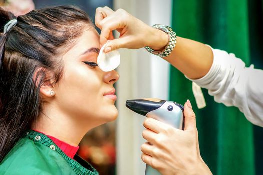 Side view of a make-up artist using aerograph making an airbrush makeup foundation on a female face in a beauty salon