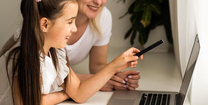 Homeschool little young girl learning online class from school teacher by digital remote internet meeting due to coronavirus pandemic. Kid looking computer and writing note with help from mother.