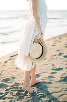 Straw hat in the hand of a barefoot woman standing on the sand. High quality photo