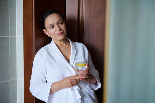 Beautiful middle-aged multi-ethnic woman in white waffle bathrobe with a glass of fresh lemon water. Pretty woman enjoys morning routine, takes care of her skin and body. Detox treatment