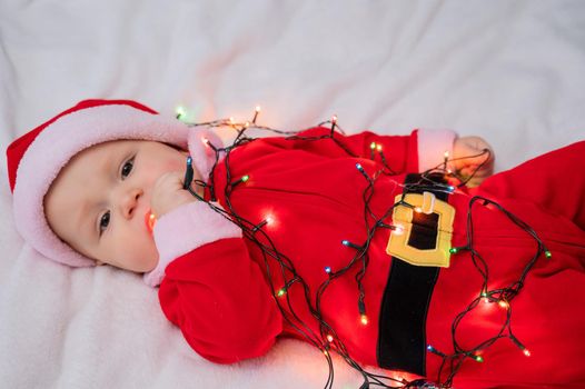 A baby boy dressed in a santa suit lies on the bed and plays with a Christmas garland