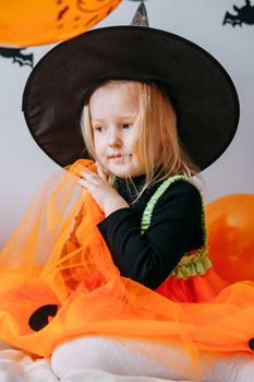 Children's Halloween - a girl in a witch hat and a carnival costume with airy orange and black balloons at home. Ready to celebrate Halloween.