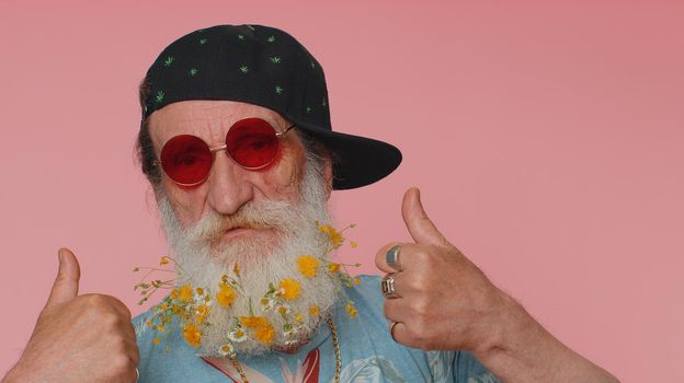 Close-up of cheerful stylish elderly man wearing sunglasses, charming smile. People emotions concept. Senior lovely mature grandfather with flowers in gray-haired beard posing isolated alone on pink
