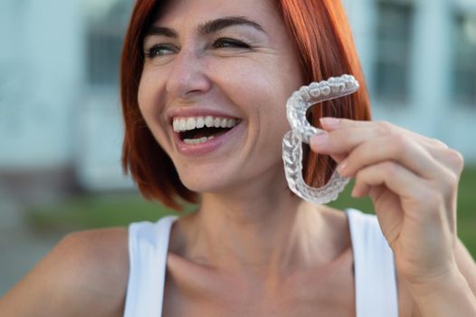Red-haired Caucasian woman holding transparent mouthguards for bite correction outdoors. A girl with a beautiful snow-white smile uses silicone braces.