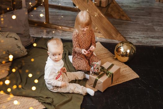 Little brother and sister play on Christmas eve in a beautiful house decorated for the New Year holidays. Children are playing with a Christmas gift. Scandinavian-style interior with live fir trees and a wooden staircase.