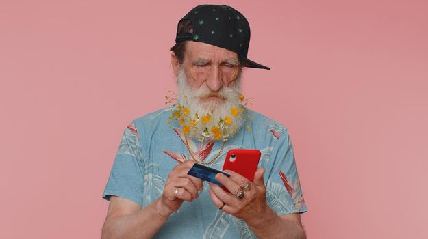 Sincere mature man customer using credit bank card and smartphone while transferring money, purchases online shopping, payment. Finance and internet. Senior grandfather alone on pink wall background