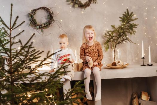 Little brother and sister play on Christmas eve in a beautiful house decorated for the New Year holidays. Children are playing with a Christmas gift. Scandinavian-style interior with live fir trees and a wooden staircase.