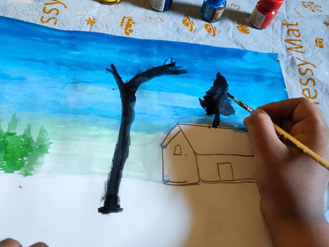 A child is painting in a paper. In the picture the child is painting a house and tree in a blue background.