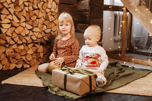 Little brother and sister play on Christmas eve in a beautiful house decorated for the New Year holidays. Children are playing with a Christmas gift. Scandinavian-style interior with live fir trees and a wooden staircase.