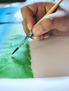 A child is painting in a paper. In the picture the child is painting grasses in a blue background.