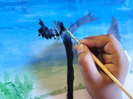 A child is painting in a paper. In the picture the child is painting a house and tree in a blue background.