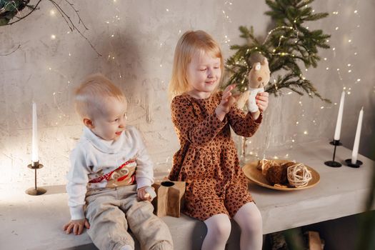 Little brother and sister play on Christmas eve in a beautiful house decorated for the New Year holidays. Children are playing with a Christmas gift. Scandinavian-style interior with live fir trees and a wooden staircase.