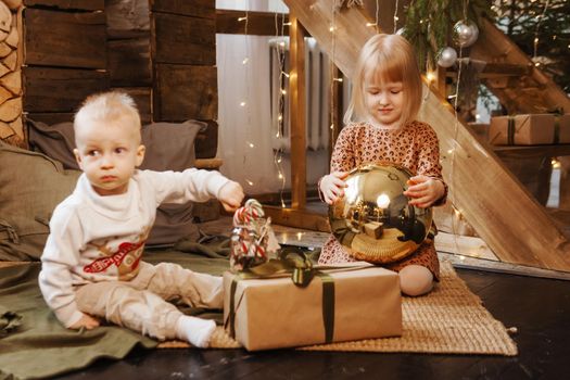 Little brother and sister play on Christmas eve in a beautiful house decorated for the New Year holidays. Children are playing with a Christmas gift. Scandinavian-style interior with live fir trees and a wooden staircase.