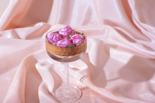 Bokal glass with roses in champagne on fabric folds background. Creative still life in pastel pink colors.