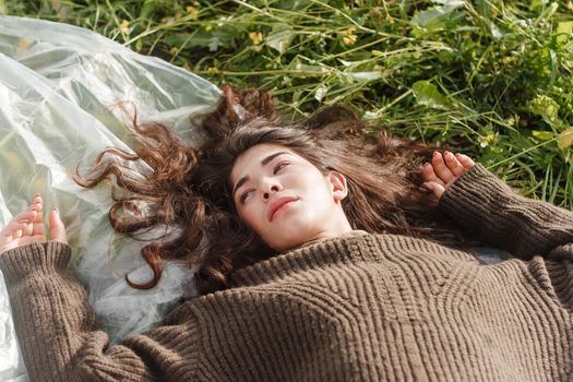 An Asian woman is lying on the ground in a field in a brown sweater. The concept of manufacturing clothing from recycled plastic. An Asian woman is lying on a plastic bag. Portrait of a dark-haired woman with loose hair.