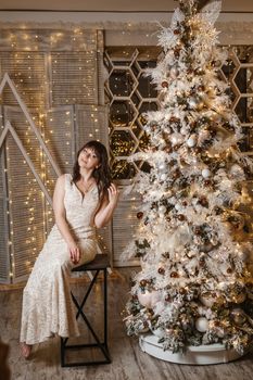 A brunette woman in a beautiful light tight dress next to a Christmas tree. The magical atmosphere of Christmas Eve on December 25. Decoration for the holiday in a light style with lots of lights and light bulbs.