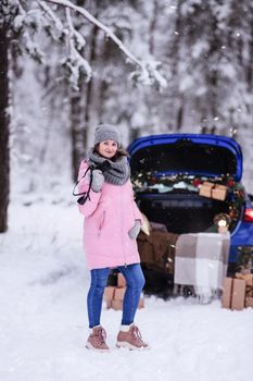 A woman in a winter snow-covered forest in the trunk of a car decorated with Christmas decor. A female photographer holds a camera in her hands.