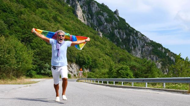 Portrait of a gray-haired senior elderly Caucasian man bisexuality with a beard and sunglasses holding a rainbow LGBTQIA flag on nature. Celebrates Pride Month, Rainbow Flag Day, gay parade