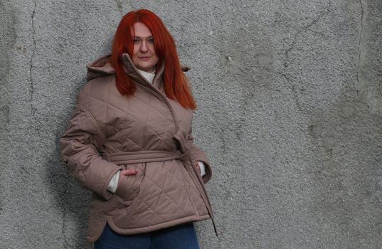 A plus-size girl with red hair in a brown jacket against an old concrete wall with her hands in her pockets.
