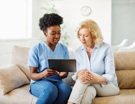 Doctor or nurse caregiver showing a tablet screen to senior woman and laughing at home or nursing home