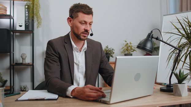 Businessman working on laptop computer wearing headset, freelance worker call center or support service operator helpline, having pleasant talk with client or colleague communication support at office