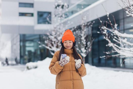 Young beautiful Asian woman listens to music from headphones while walking on a winter snowy day, uses an online application for the phone