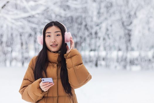 Beautiful Japanese teenager walks in the winter park uses the phone and listens to music with cute pink headphones