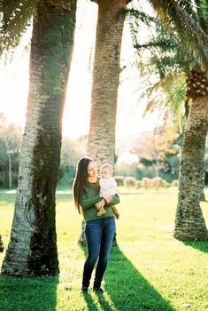 Mom looks at the baby in her arms while standing under a large palm tree. High quality photo