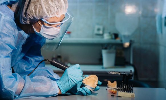 A masked and gloved dental technician works on a prosthetic tooth in his lab.