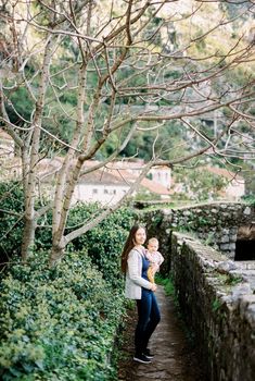 Mom with a baby stands on a hill near a stone fence overlooking old houses. High quality photo