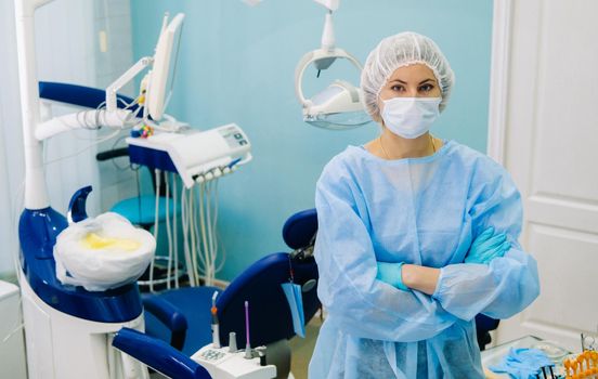 a female dentist wearing a medical mask and rubber gloves poses for the camera and folds her arms in her office.