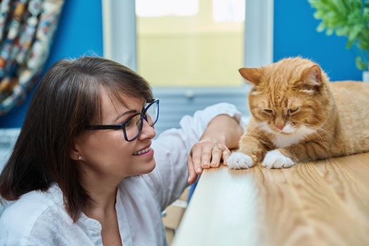 Middle aged woman talking with ginger pet cat, home interior background. Friendship, love, care, leisure, lifestyle, animals people house concept