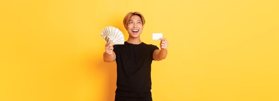 Happy good-looking asian guy looking thoughtful and pleased upper left corner while showing money and credit card, yellow background.