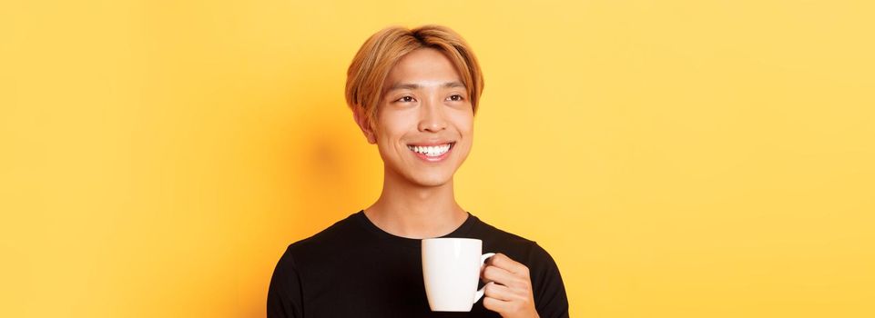 Close-up of happy handsome young asian guy with blond hair, looking dreamy and smiling while drinking coffee or tea, standing over yellow background.