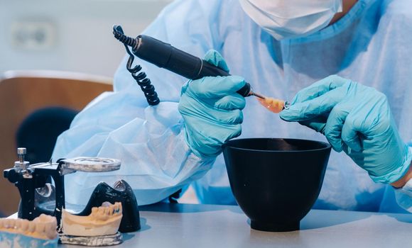 A masked and gloved dental technician works on a prosthetic tooth in his lab.