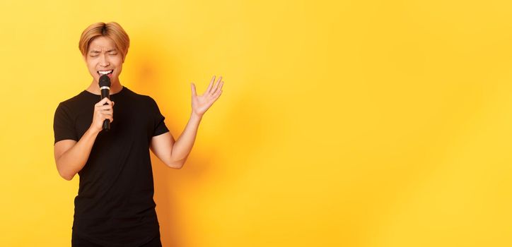 Handsome asian male singer, korean guy singing song at karaoke in microphone with passion, standing over yellow background.