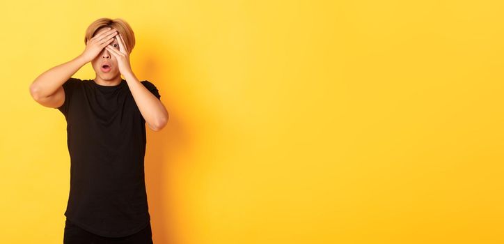 Portrait of embarrassed asian guy with blond hair, gasping startled and shut eyes, peeking through fingers, yellow background.