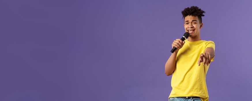 This song is for you. Portrait of romantic carefree hispanic man singing karaoke, pointing at camera as dedicate his performance, holding microphone, standing purple background.
