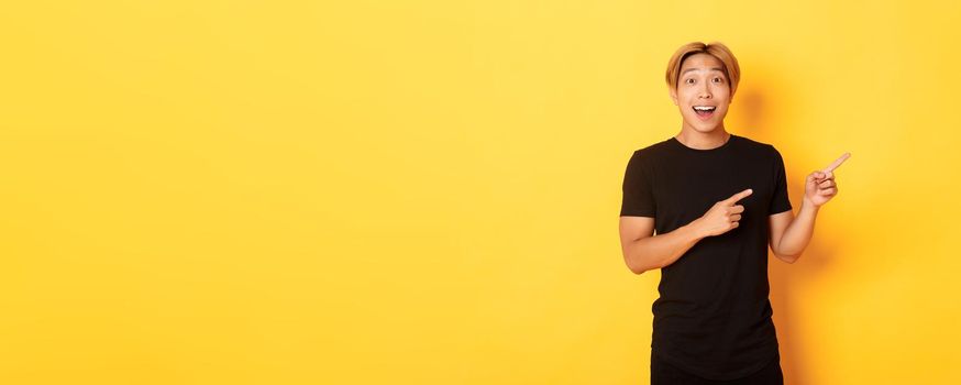 Amazed handsome korean guy, smiling fascinated and pointing fingers right, showing way, standing yellow background.