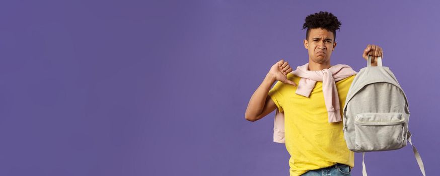 Education, university and trends concept. Portrait of gloomy disappointed young male student complaining on ugly new backpack, show thumbs-down and grimacing displeased, purple background.