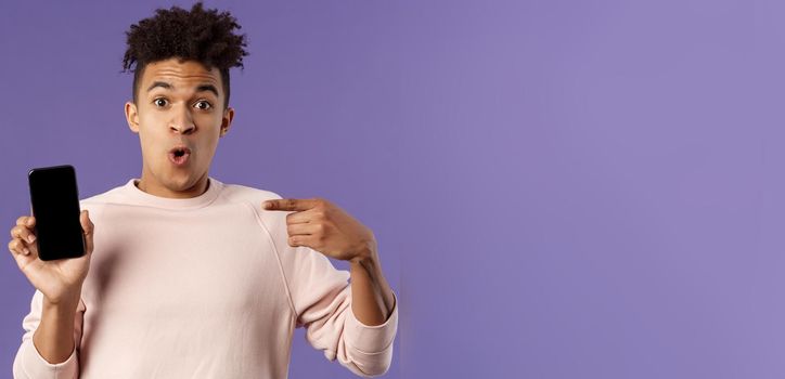 Close-up portrait of excited, cheerful young hispanic male geek talking about his device, new gadget or application, holding mobile phone, pointing at smartphone with amused face.