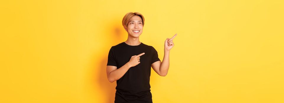 Portrait of handsome korean blond guy, pointing fingers upper left corner and looking satisfied with pleased smile, yellow background.