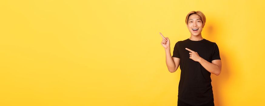 Portrait of cheerful asian man pointing fingers upper left corner, smiling and showing banner satisfied, yellow background.