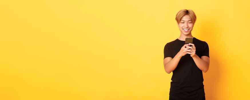 Portrait of handsome stylish asian guy with blond hair, using mobile phone and smiling, yellow background.