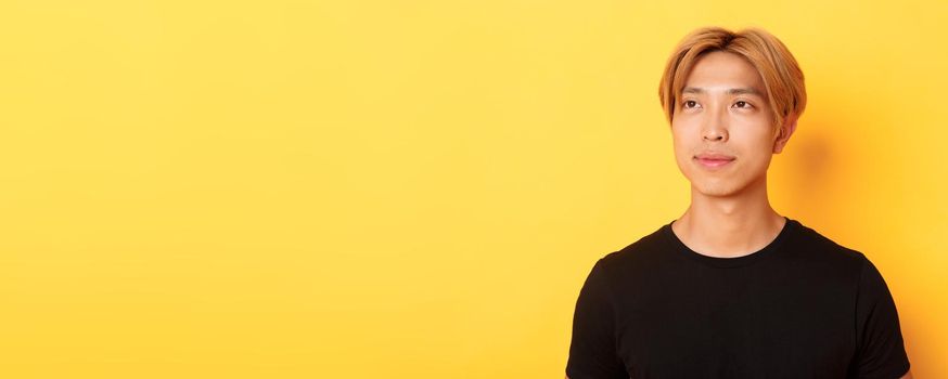 Close-up of handsome stylish korean guy with fair hair, looking dreamy and hopeful upper left corner, smiling pleased, standing yellow background.